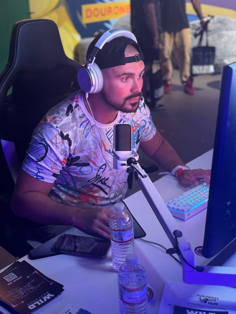 Man wearing purple headphones and patterned shirt sitting at gaming desk with microphone and monitor
